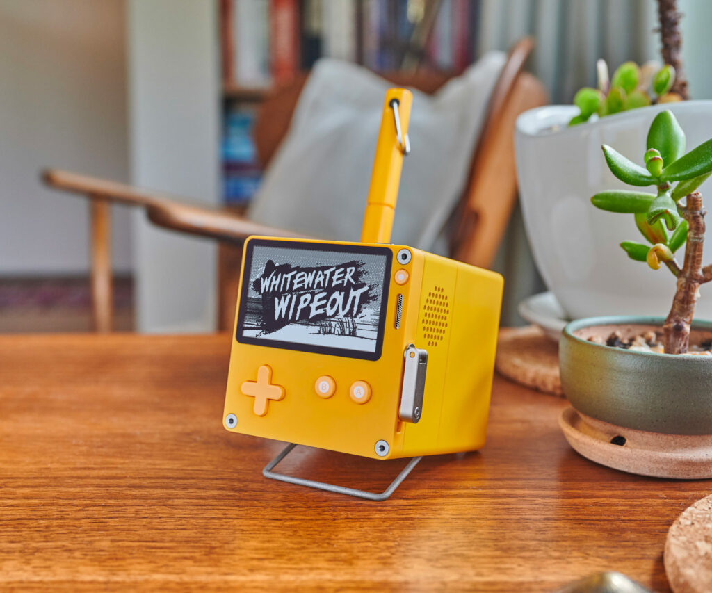A Playdate attached to a stereo dock sits on a side table in a living room, next to a potted succulent. The Playdate screen is displaying the title screen from the game Whitewater Wipeout. The yellow pen included with the dock rests in the pen holder.