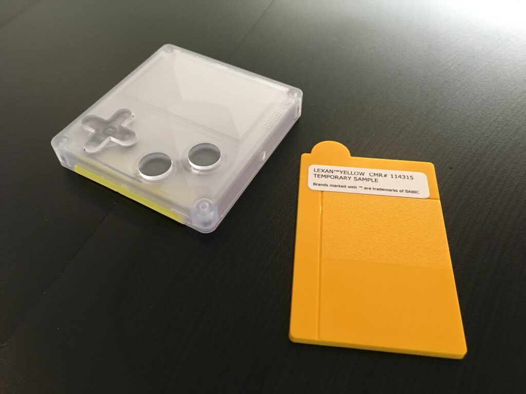 A transparent plastic Playdate case next to a rectangular piece of yellow plastic with a sticker reading Lexan yellow CMR# 114315 Temporary Sample