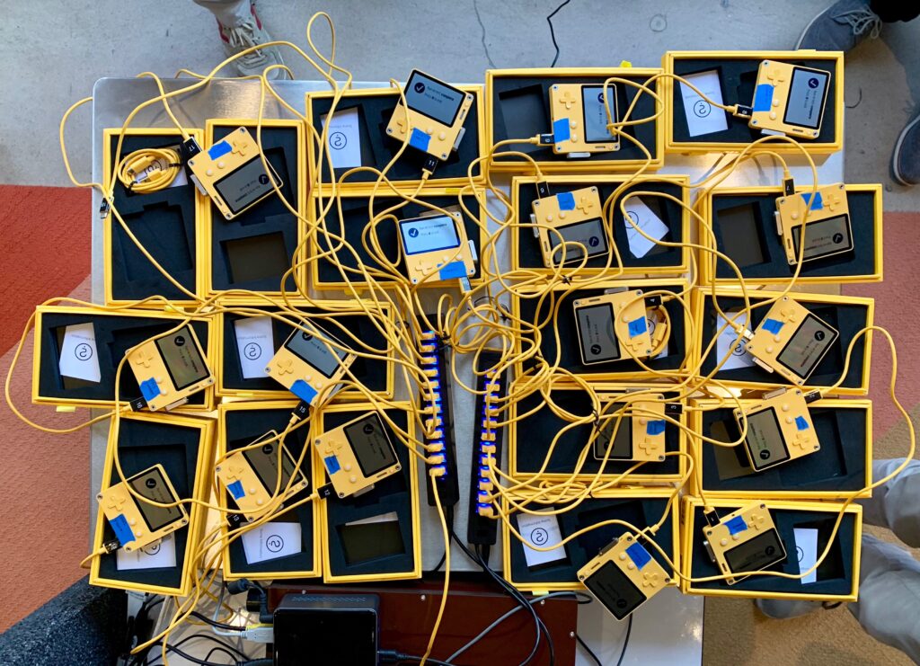 Many Playdate units marked with blue tape are connected via USB cords to two large USB hubs